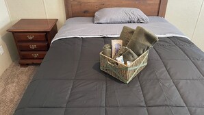 Una cama bien hecha con colcha oscura, cabecera de madera y una pequeña cesta de mimbre sobre la cama con toallas y una caja de pañuelos.