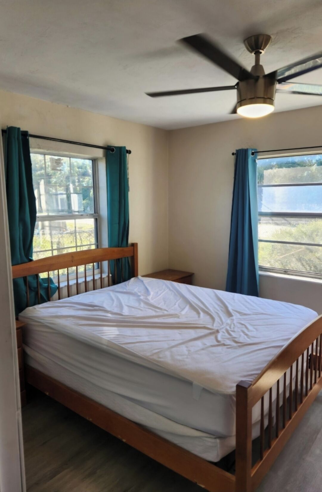 Una cama individual con estructura de madera en una habitación con ventilador de techo y dos ventanas con cortinas.