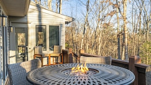 Um deck com uma mesa redonda e cadeiras, um poço de fogo e vista para árvores.
