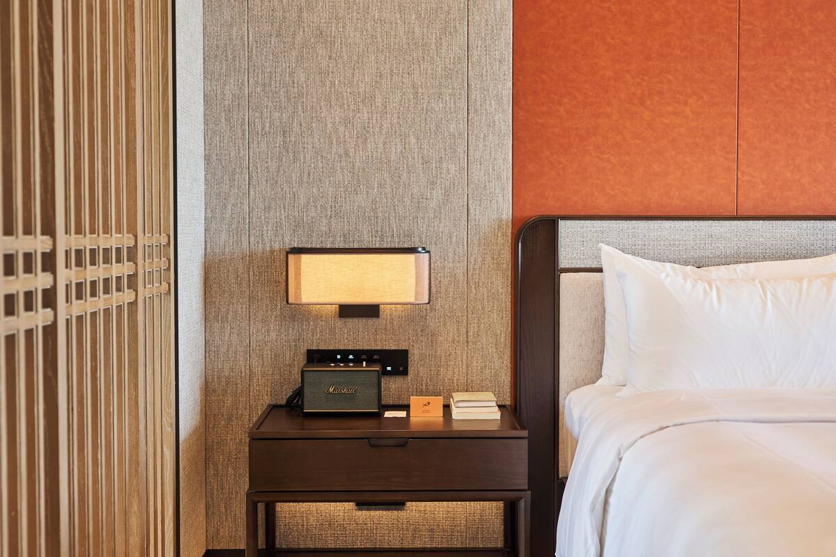 Ein ordentlich eingerichtetes Hotelzimmer mit Nachttisch aus Holz, einer Lampe, einem Radio und einem Bett mit weißer Bettwäsche.