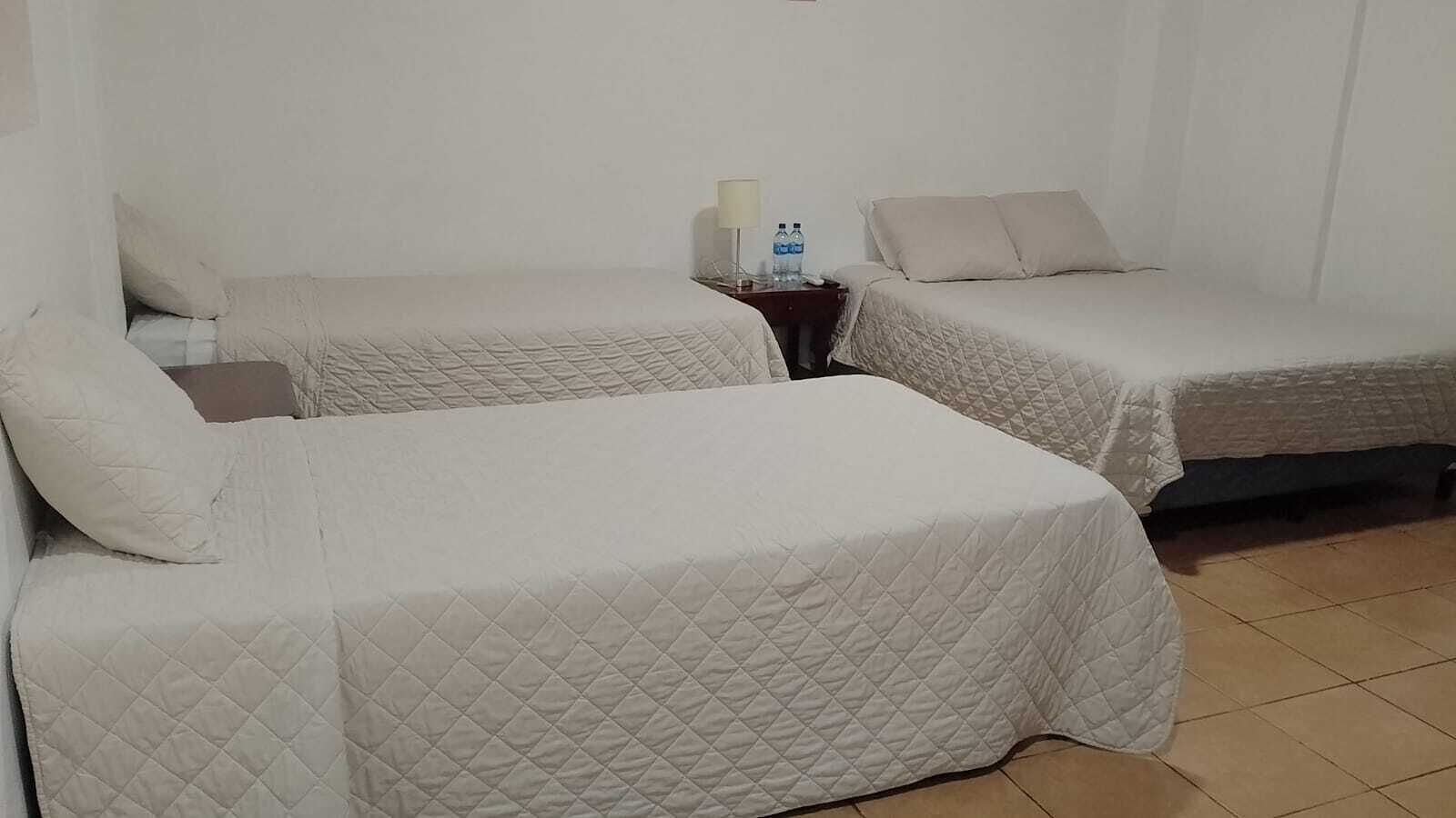 A hotel room with two beds, each with a quilted bedspread and pillows. There is a nightstand with a lamp and a bottle of water.