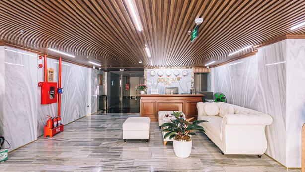 Un vestíbulo de hotel con techo de madera, paredes de mármol, un mostrador de recepción, un sofá blanco y una planta en maceta.