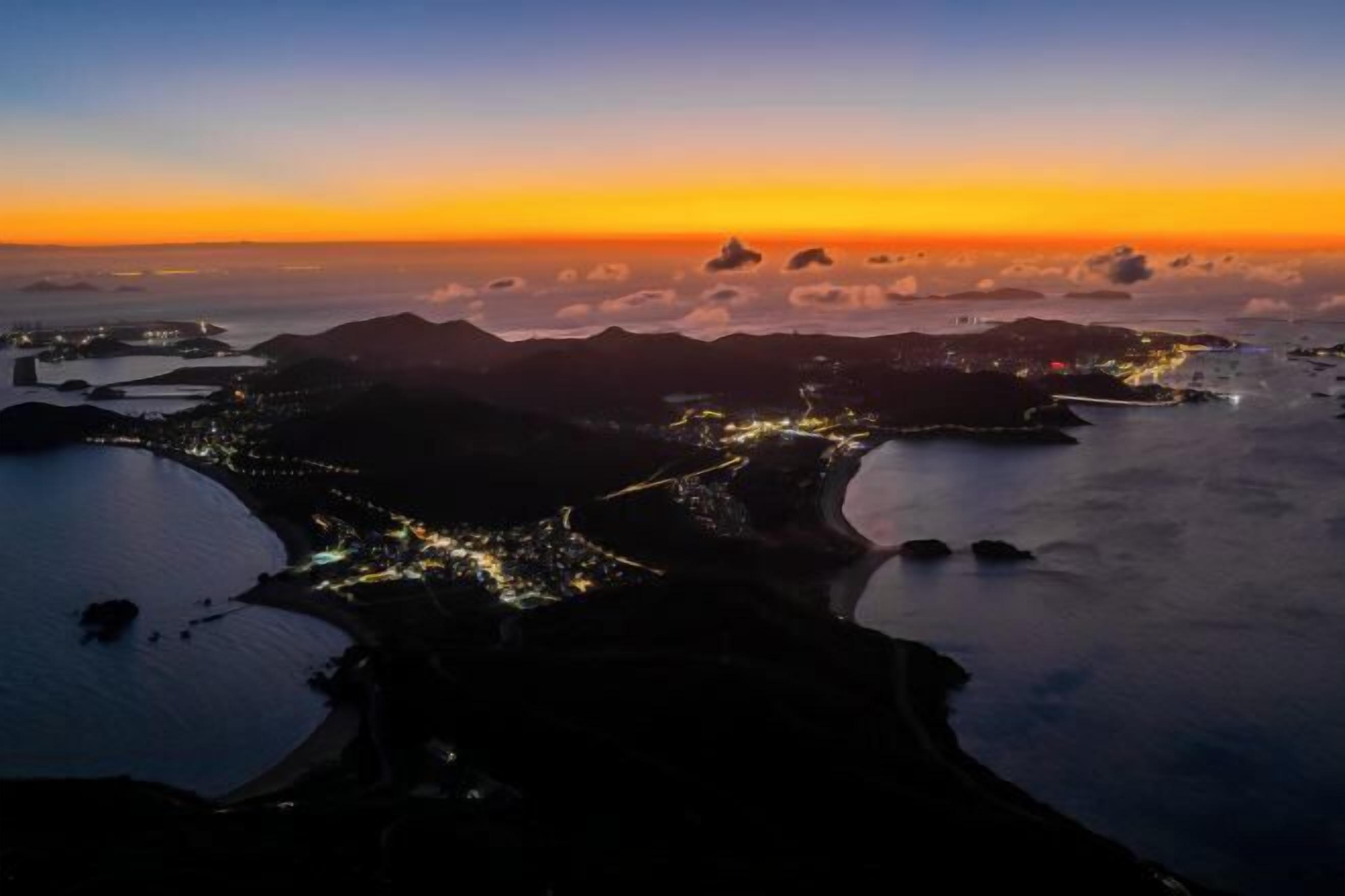 Vue aérienne d’une ville côtière au coucher du soleil, avec des rues illuminées et des étendues d’eau.