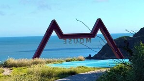 A red 'M' shaped sign with the word 'MUSE' on a grassy area with a view of the ocean and a pool.