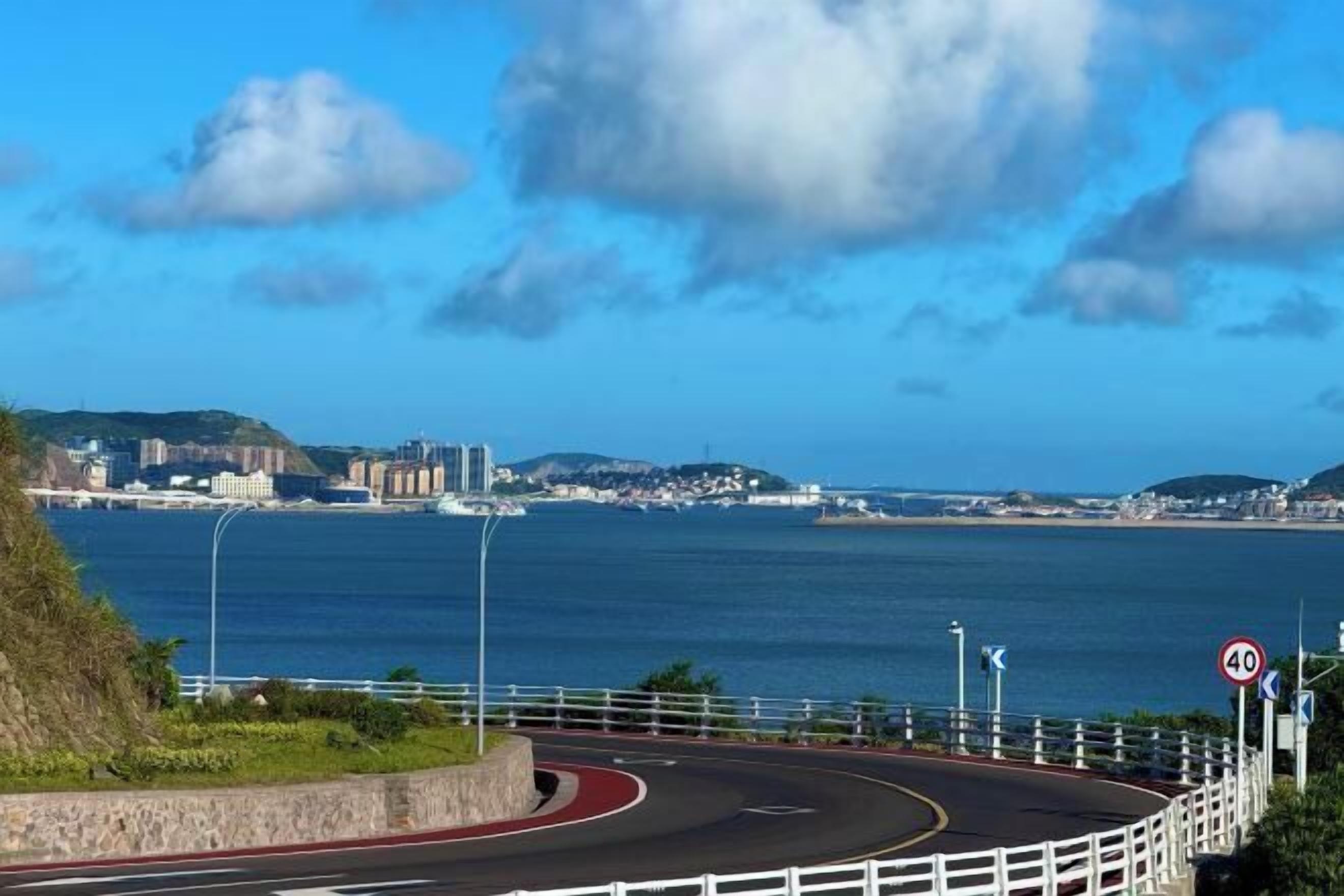 Carretera costera con un cartel de límite de velocidad de 40, una rotonda y vista de un paisaje urbano al otro lado del agua.