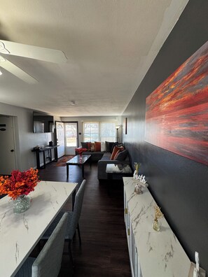 A modern living room with a large painting, a dining area with a marble table, and a kitchen in the background.