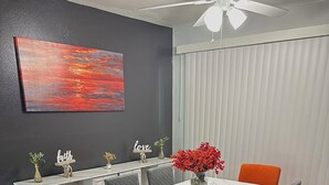 Un coin repas avec une table blanche, des chaises grises et orange, un meuble en marbre et un ventilateur de plafond.