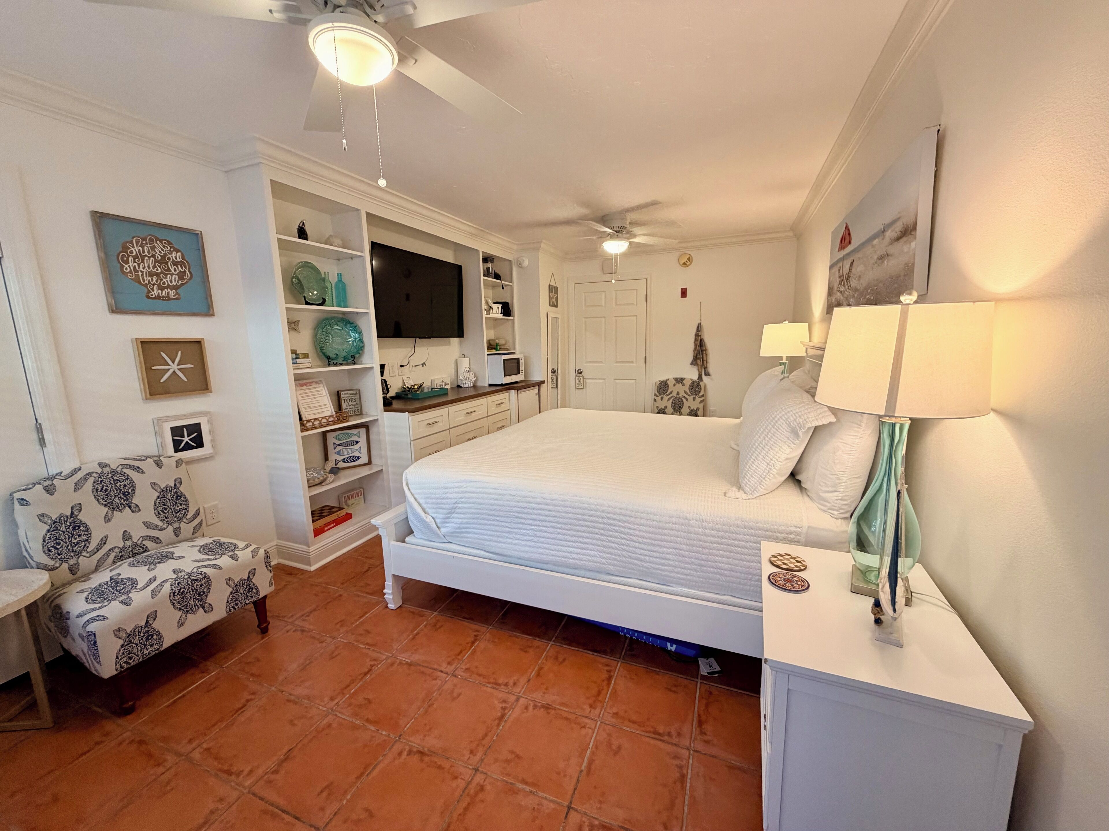 A bedroom with a bed, a chair, a vanity, a TV, and a ceiling fan.