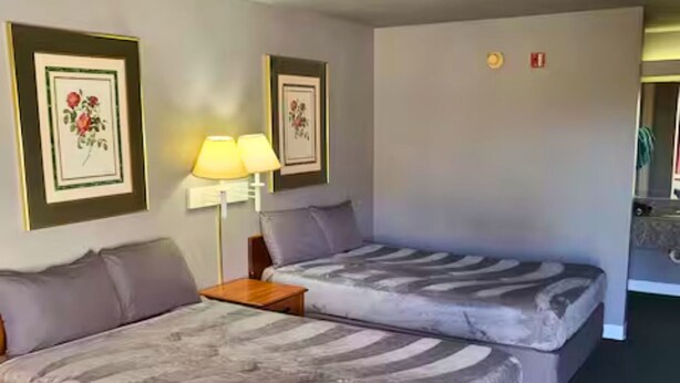 A hotel room with two beds, a nightstand with lamps, framed floral artwork on the walls, and a visible fire alarm. - Room 124 - 2 Double Beds (Livingston)