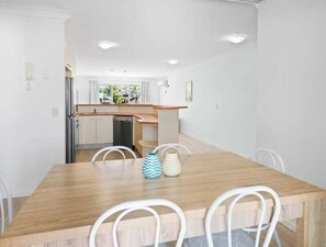 A dining area with a wooden table and white chairs, a kitchen in the background, and a view of greenery outside. - Mermaid Beach Resort Stay + 2BR + Balcony & Pool (Gold Coast)