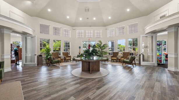 A spacious lobby with a central wooden table, surrounded by chairs, large windows, and a high ceiling.