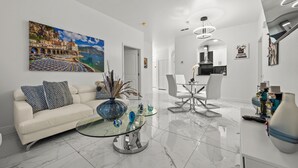 A modern living room with a white sofa, a glass coffee table, and a large painting of a coastal scene.