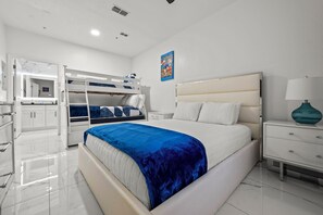 Une chambre moderne avec un lit superposé, un grand lit recouvert d’une couverture bleue, des meubles blancs et une affiche encadrée accrochée au mur.