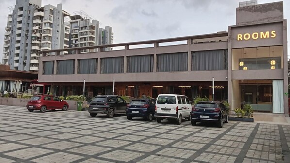 A hotel with a parking lot containing several cars. The building has a sign that reads "ROOMS" and features a modern architectural style with large windows and a flat roof. - Hotel Euphoria The Fine Dine (Surat)