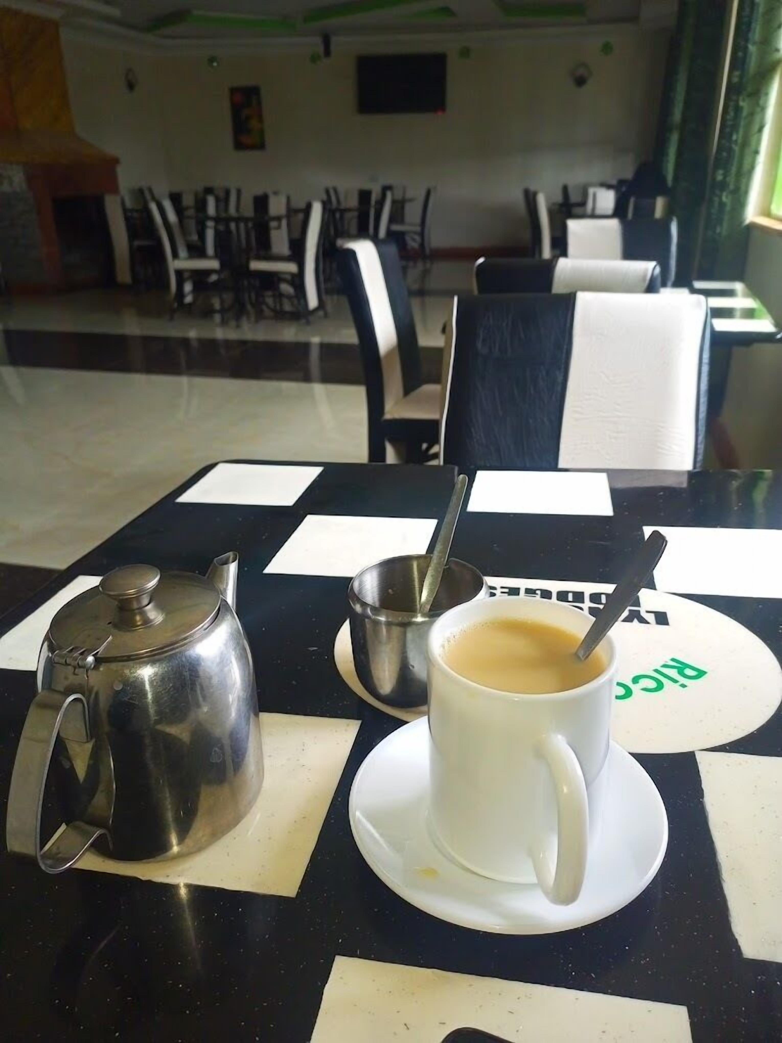 Una mesa con un hervidor de agua, una taza de café y un pequeño recipiente metálico con una cuchara. La mesa está en un comedor con sillas y mesas al fondo.