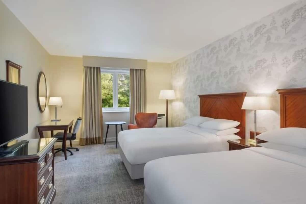A hotel room with two beds, a TV, a desk, a chair, and a window with curtains.