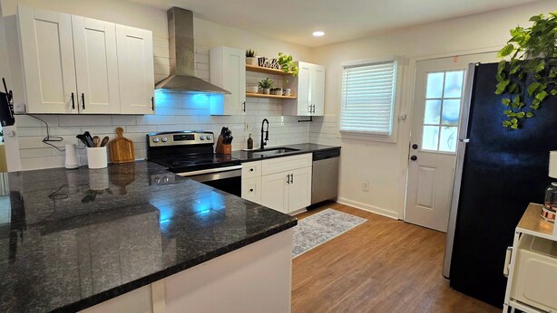 A modern kitchen with white cabinets, a black countertop, and a stainless steel range hood. There are plants on the shelves and a refrigerator on the right. - Cozy & Stylish Home near Atlanta (Forest Park)