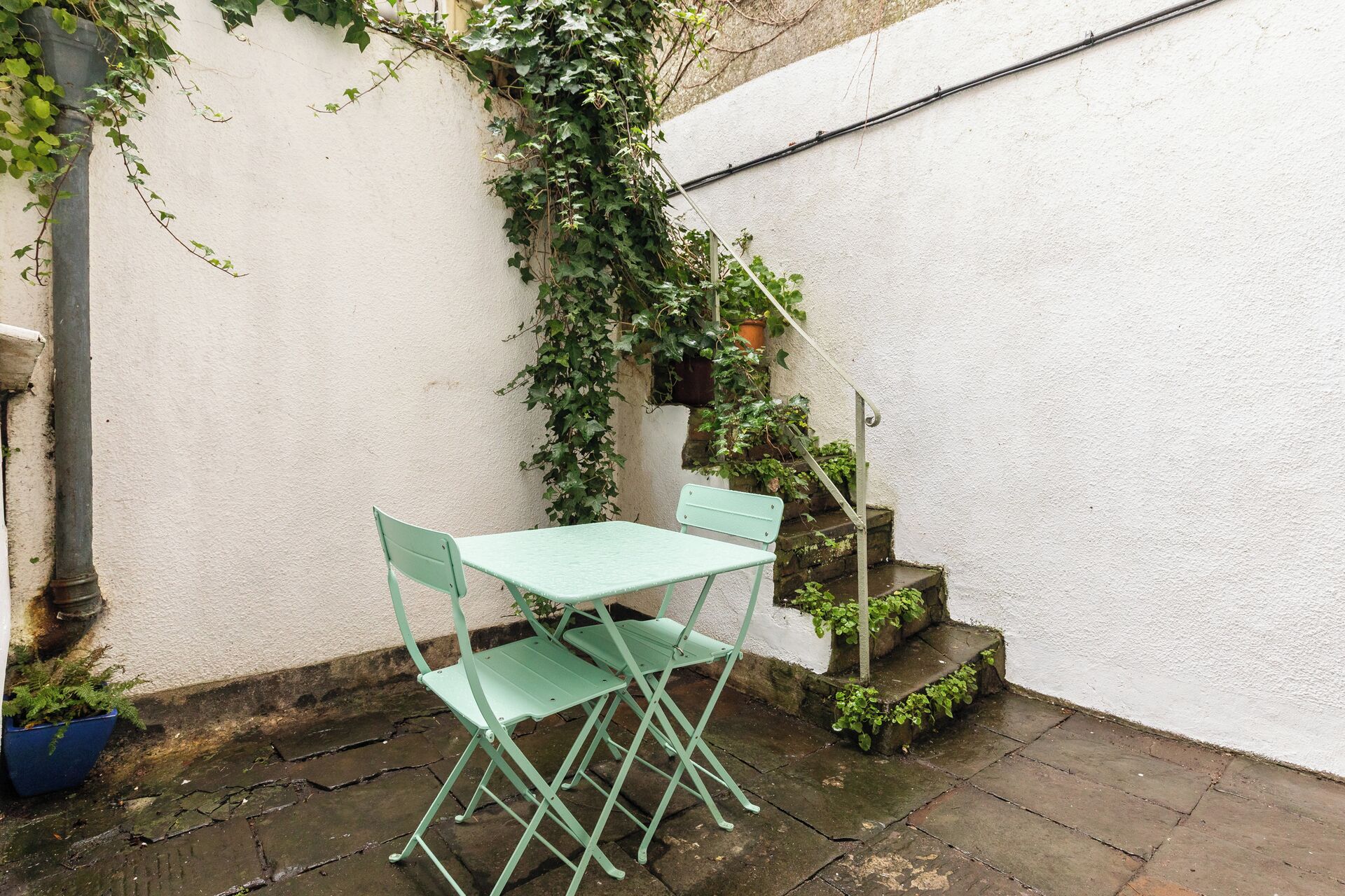 A small outdoor seating area with a table and two chairs, surrounded by a white wall and covered with ivy.