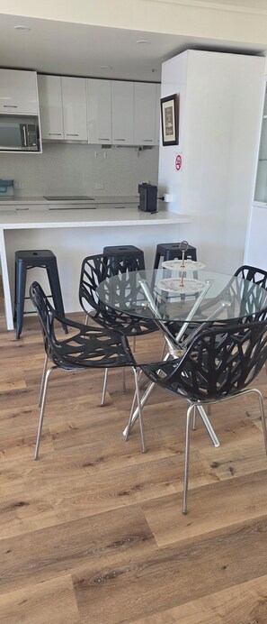 A modern kitchen with white cabinets, a microwave, and a coffee machine. There is a dining area with a glass table and black chairs. - Bright 2 b/room apartment -up to 6 guests-parking incl. Central to everything. (Melbourne)
