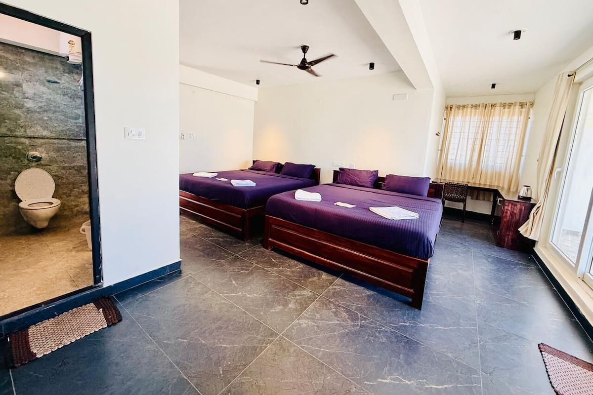 A hotel room with two beds, a ceiling fan, a desk, a window with curtains, and a bathroom with a toilet and shower.