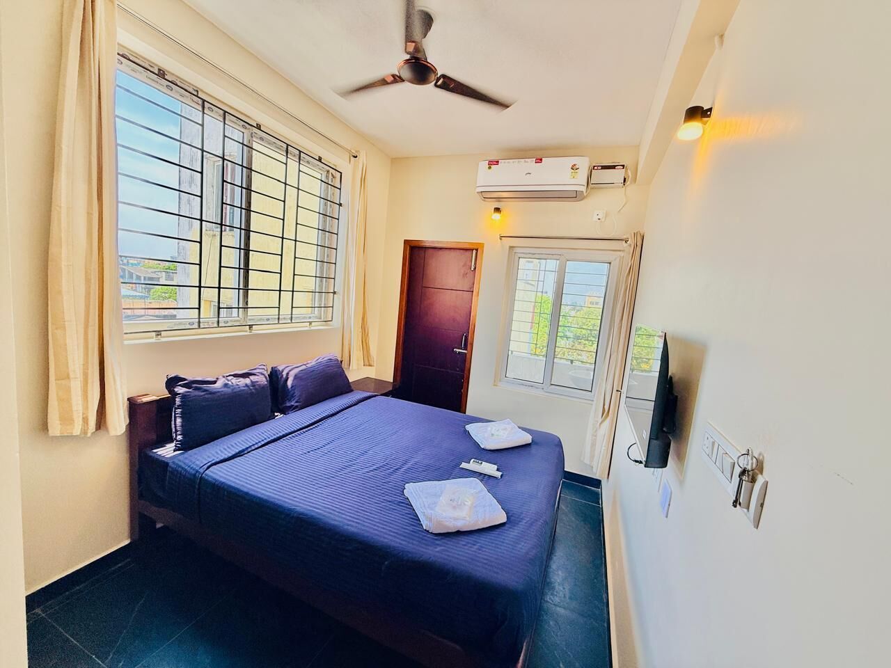 A single bed with a dark blue bedspread, a ceiling fan, an air conditioning unit, and a television on the wall.