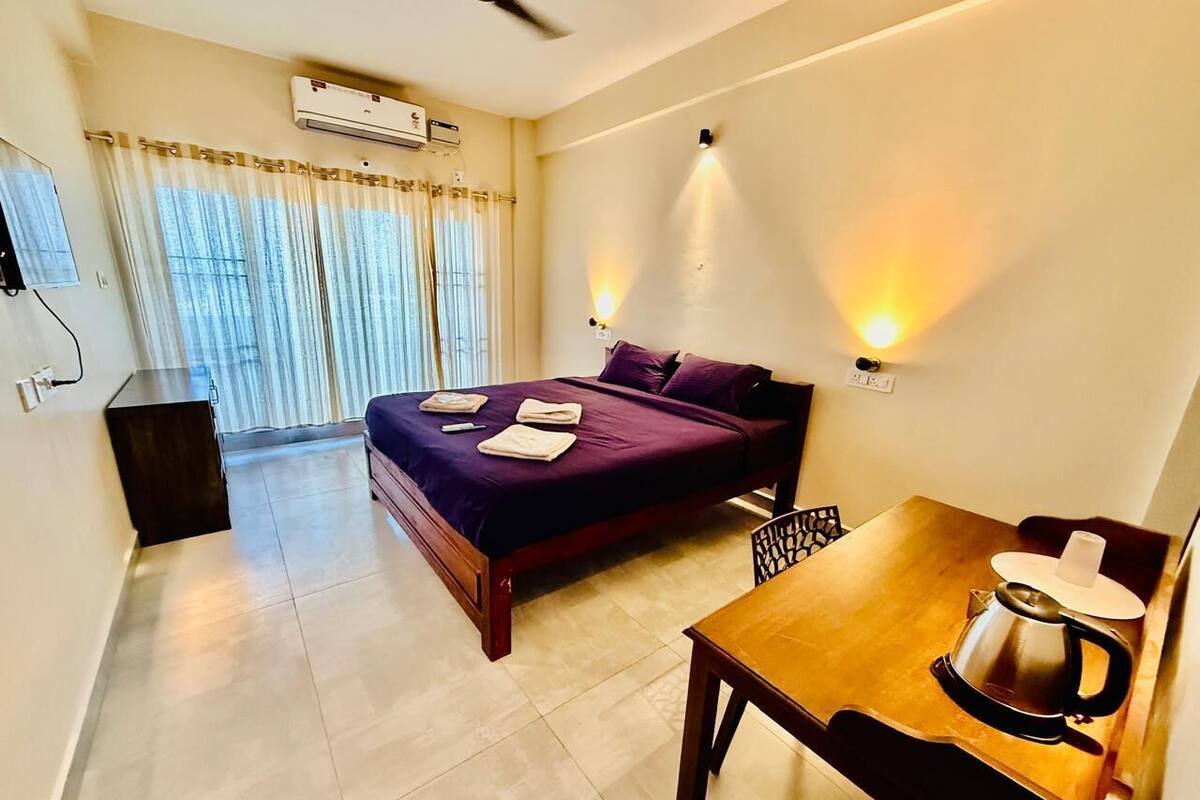 A hotel room with a bed, a table with a kettle, a ceiling fan, and a window with curtains.