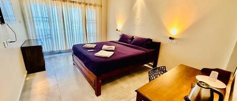 A hotel room with a bed, a table with a kettle, a ceiling fan, and a window with curtains.