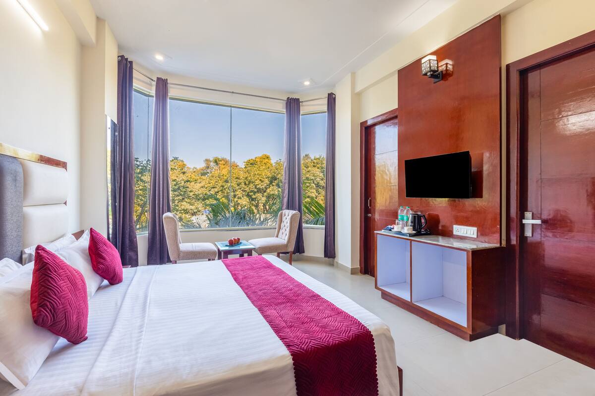 A hotel room with a large bed, a TV, a desk with a coffee maker, and a view of trees outside.