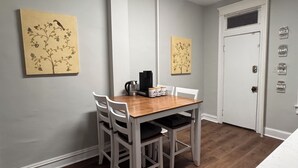 Ein Essbereich mit Holztisch und weißen Stühlen, zwei an der Wand befestigten Bildern, einer Kaffeemaschine und einer Tür mit Aufklebern.