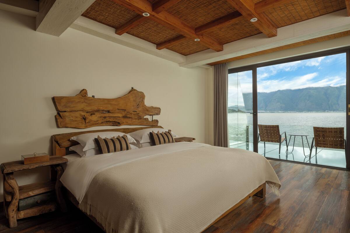 A large bed with a wooden headboard, a nightstand, and a view of a lake through large windows.