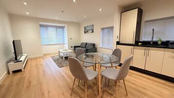 A modern living space with a dining area, a sofa, a TV, and a kitchen area with cabinets and a window.