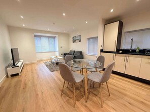 A modern living room with a glass dining table, grey chairs, a flat-screen TV, and a sofa. - Sleek Modern 2BDR Flat in Charming Pinner (Greater London)