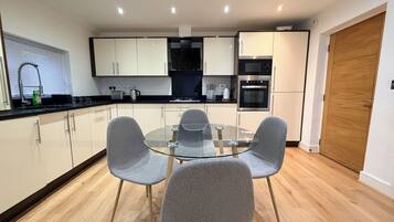 A modern kitchen with a glass dining table and grey chairs, wooden flooring, and built-in appliances.