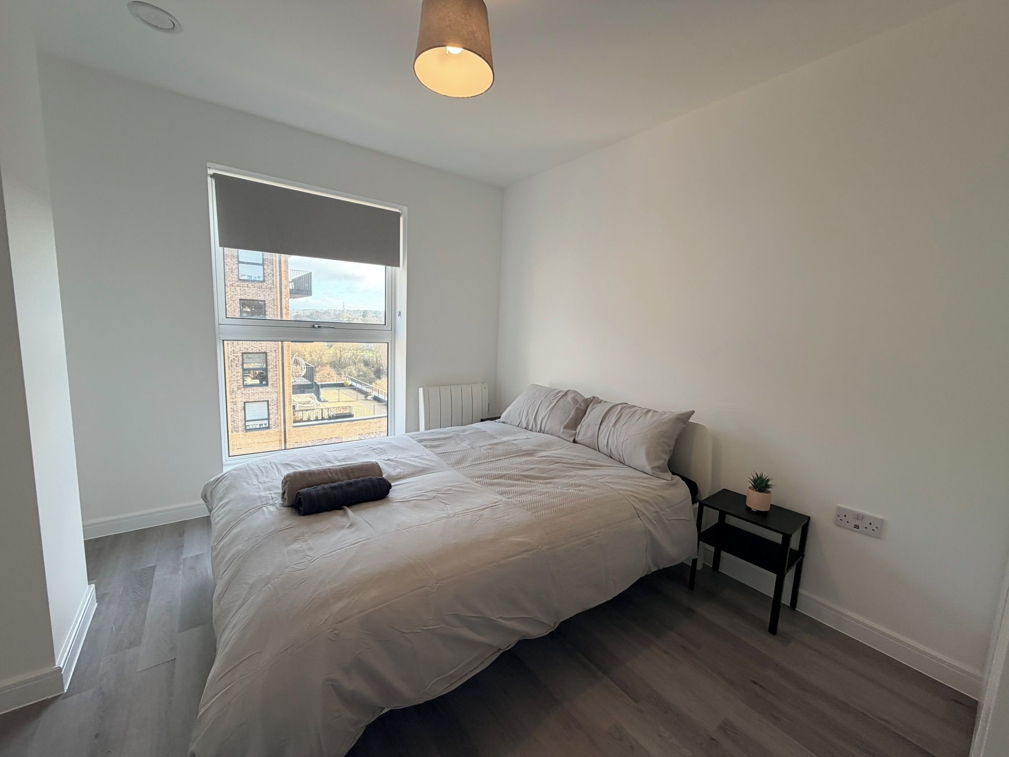 A bedroom with a large bed, a bedside table with a plant, and a window with a view of buildings.