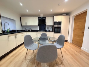 A modern kitchen with a glass dining table and grey chairs, wooden flooring, and built-in appliances. - Sleek Modern 2BDR Flat in Charming Pinner (Greater London)