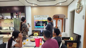 Un grupo de personas en una habitación con una televisión mostrando un programa, una mesa con un cuaderno y un cojín alegre.