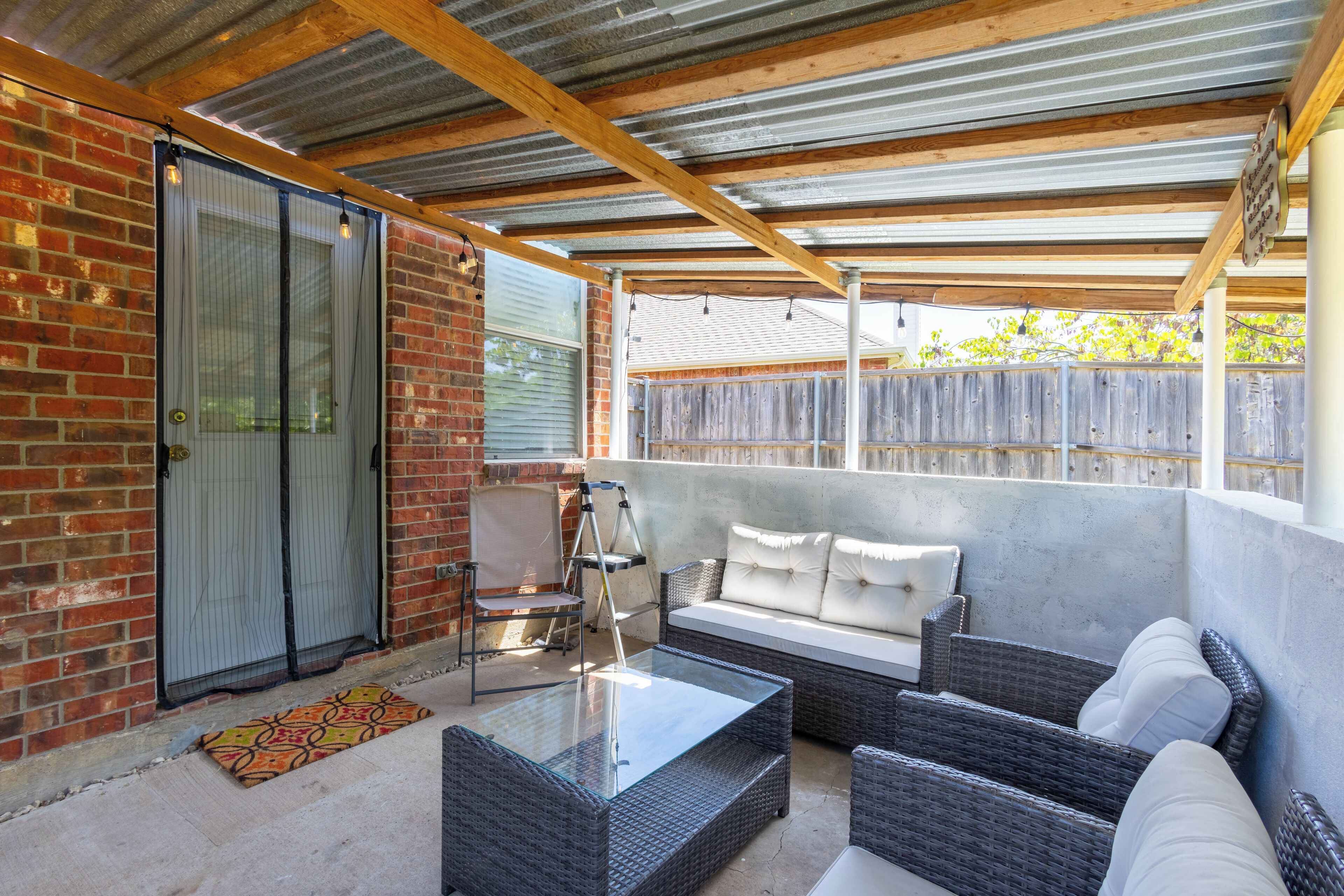 A covered patio area with wicker furniture, a glass coffee table, and a brick wall.