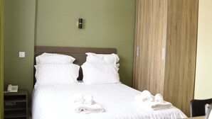 Een net opgemaakt bed met witte lakens, een houten garderobe en een nachtkastje met een telefoon en een waterkoker.