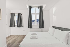 A bedroom with a large bed, a window with curtains, a radiator, and a view of buildings outside.