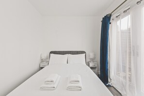 A neatly made bed with two folded white towels, a bedside table with a lamp, and a window with blue curtains. - Beautiful 4BDR Townhouse with Free Parking (Hayes)