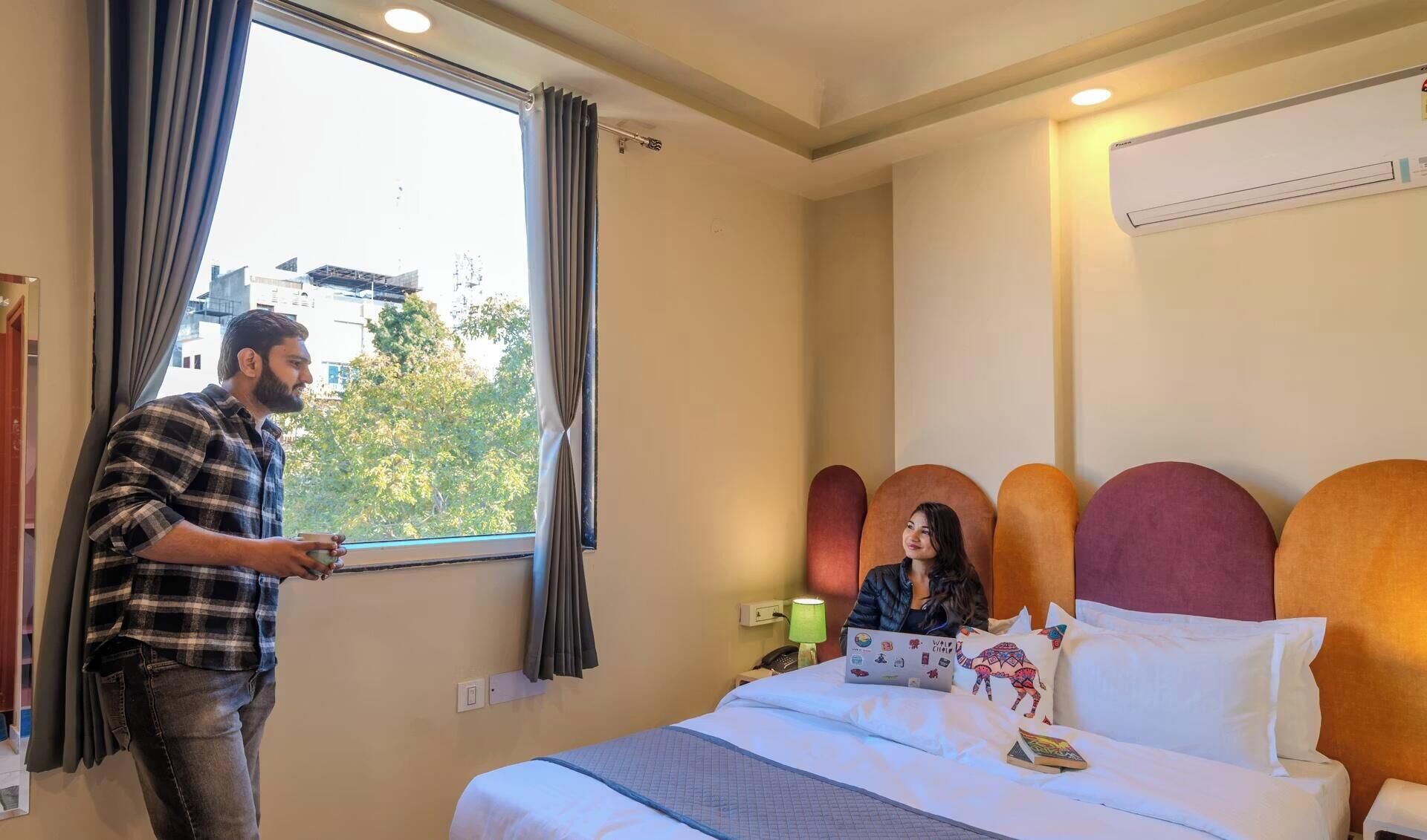 Een hotelkamer met een bed, een raam met gordijnen, een luchtverkoeler aan de muur en een bedlampje.