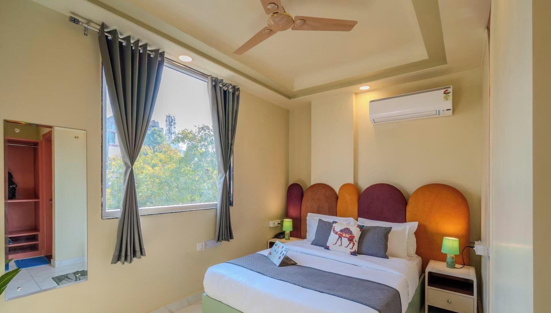 Habitación de hotel con cama, mesita de noche, ventilador de techo, ventana con cortinas y unidad de aire acondicionado.