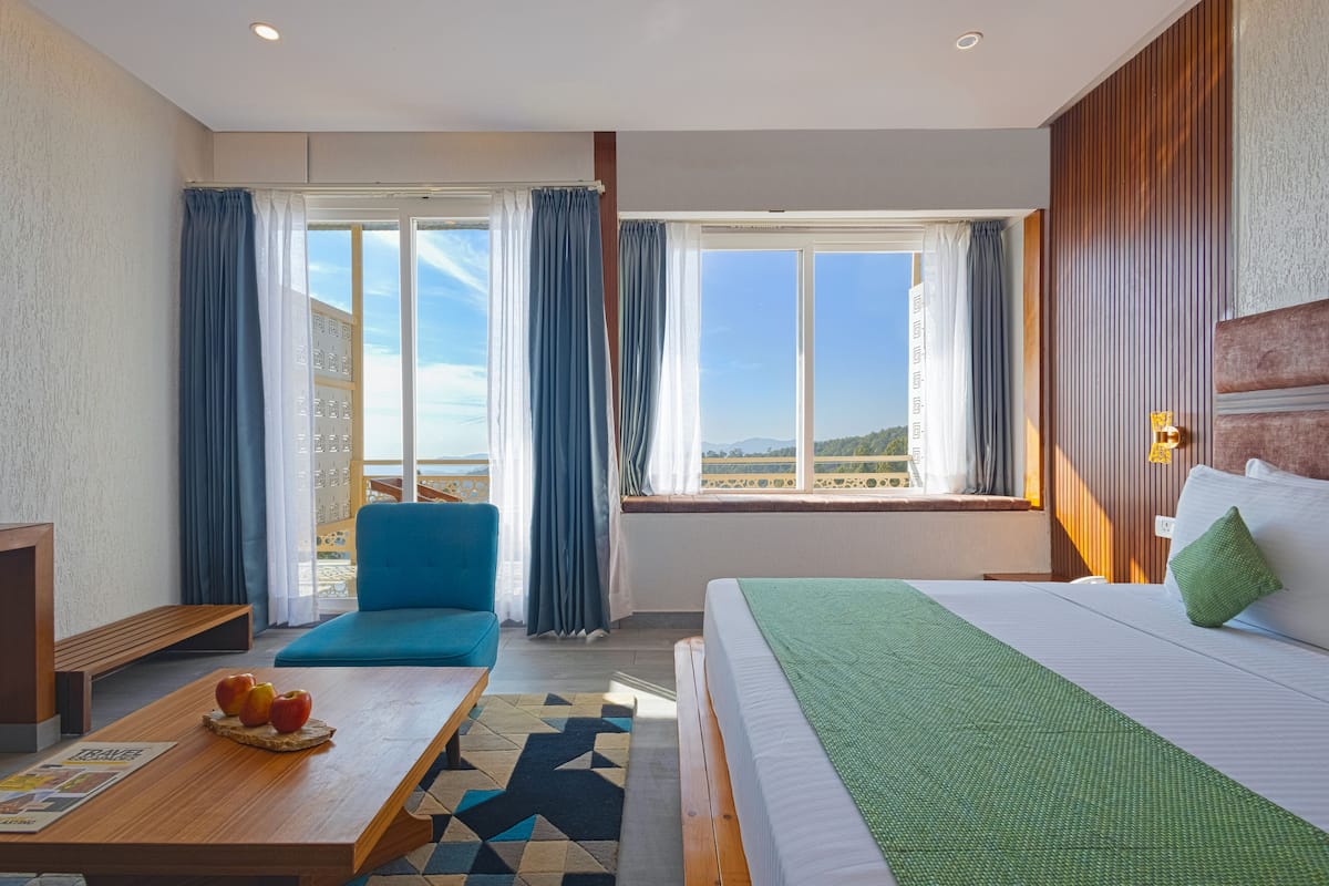A hotel room with a bed, a sofa, a wooden table with fruit, and a large window with a view.