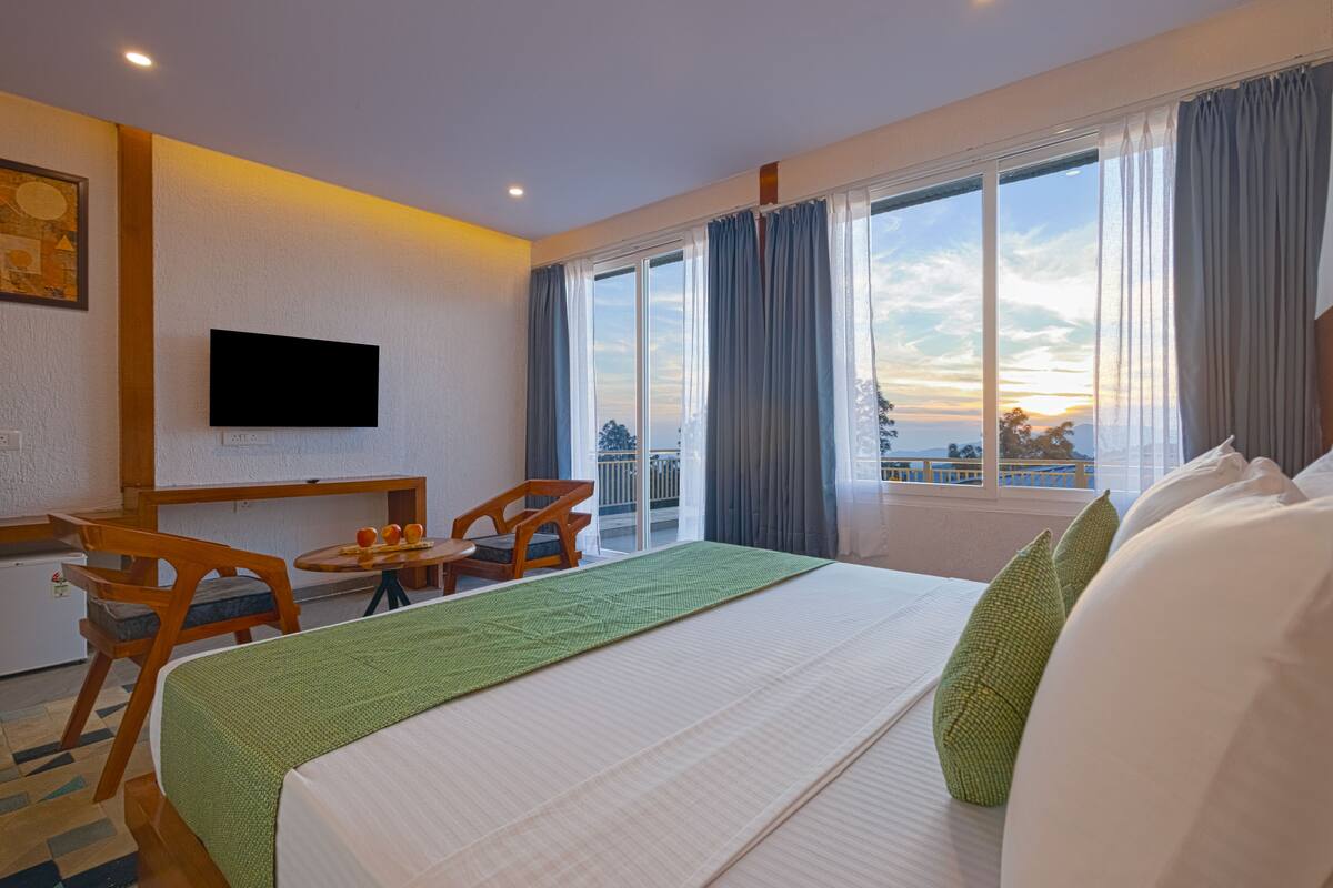 A hotel room with a bed, a TV mounted on the wall, a balcony with curtains, and a small table with fruit.