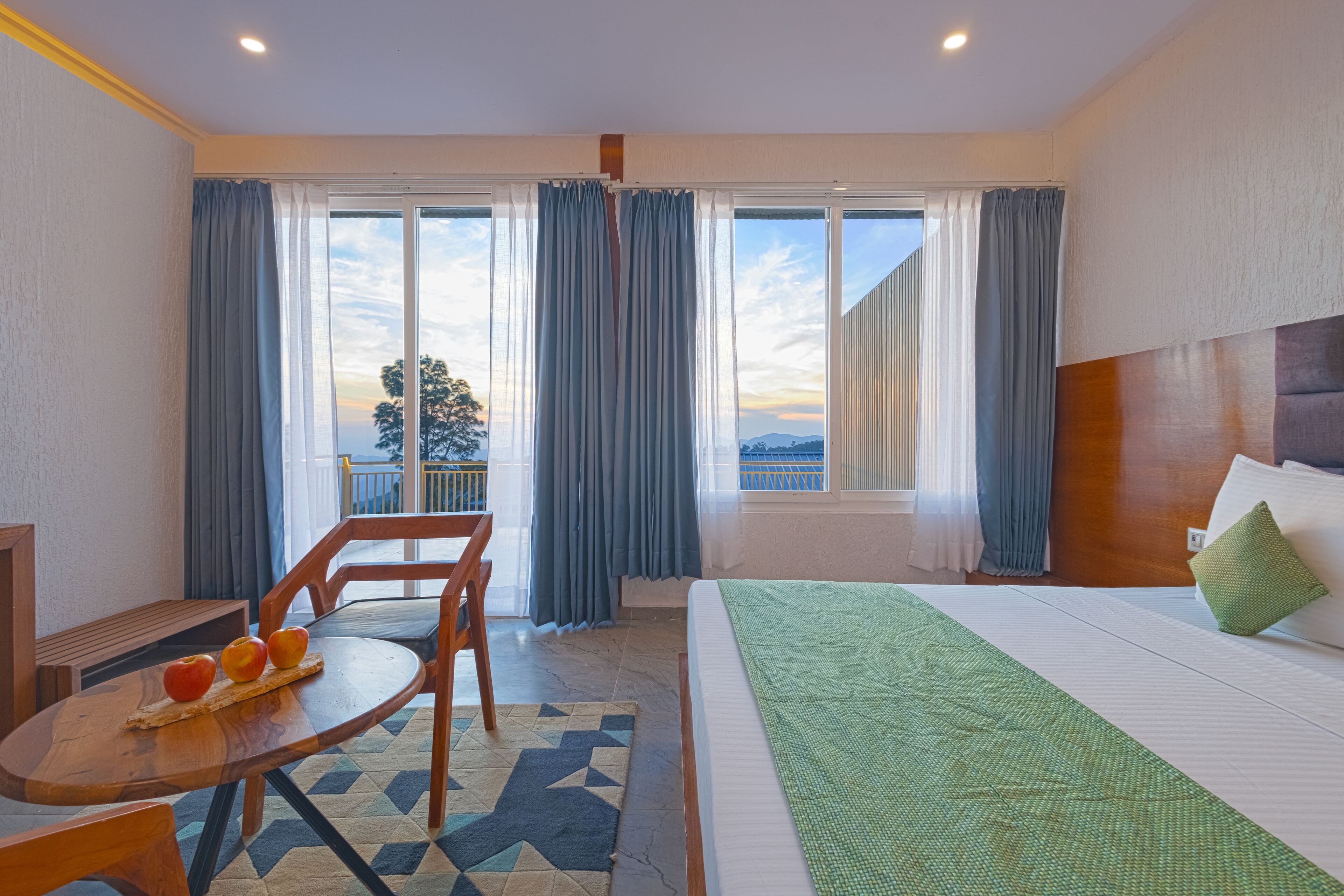 Habitación de hotel con cama, una mesa con frutas, una silla y vistas al exterior a través de amplios ventanales.