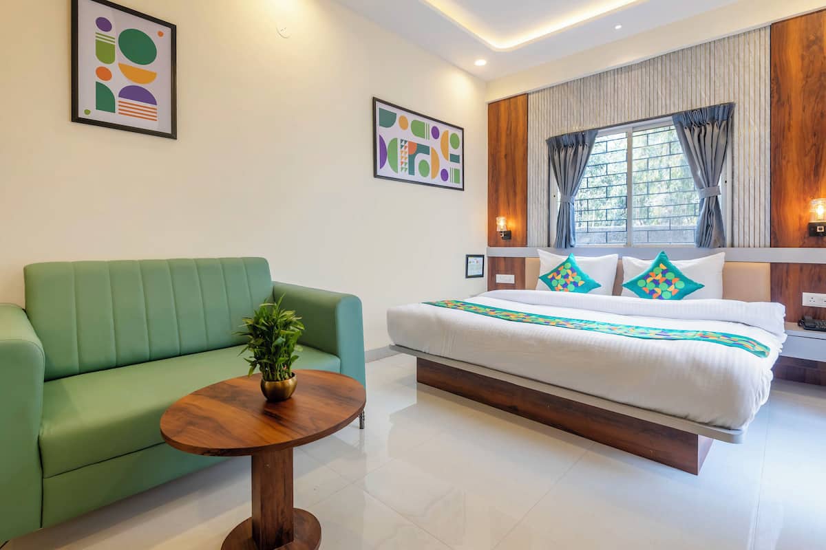 Une chambre d’hôtel moderne avec un canapé vert, une table basse en bois, un grand lit recouvert d’une couette blanche et agrémenté de coussins colorés, ainsi que deux tableaux abstraits accrochés aux murs.