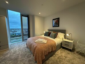 A bedroom with a bed, bedside tables, a nightstand, a window with a view, and a door. - Victoria Riverside Retreat | 2BR Retreat (Greater Manchester)