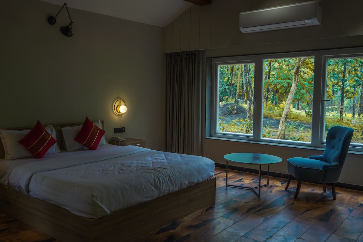 Ein Schlafzimmer mit einem großen Bett, Nachttischlampen, einem Nachttisch, einem kleinen Tisch, einem Sessel und einem Fenster mit Blick auf Bäume.