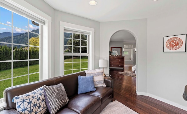 A living room with a leather sofa, patterned cushions, a lamp, and a view of a green landscape through large windows. - The Tyrolean: Pool, Hot Tub & Ski Hill Charm (Leavenworth)