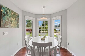 A dining area with a round table, white chairs, and a pendant light. There are three windows with white frames, and a painting is visible on the wall. - The Tyrolean: Pool, Hot Tub & Ski Hill Charm (Leavenworth)